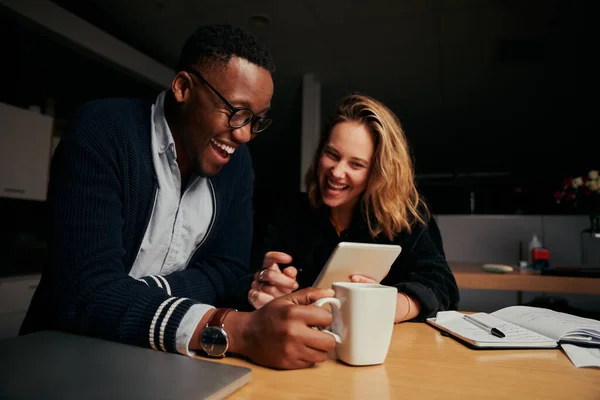 Feliz hombre de negocios y mujer de negocios sentados juntos riendo mientras miran la tableta digital durante el descanso por la noche - dos colegas sonriendo y trabajando hasta tarde juntos — Foto de Stock