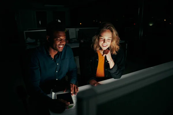 Twee lachende jonge multi-etnische zakencollega 's die' s avonds laat aan een bureau in het donker op de computer zitten te werken - twee collega 's die maandenlang de deadlines afronden — Stockfoto