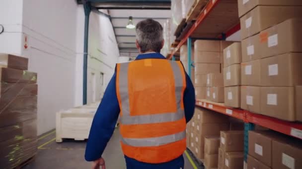 Prise de vue de suivi d'un employé de l'entrepôt arrière dans un gilet de protection enlève le casque de sécurité de la tête tout en marchant dans la salle d'entreposage — Video