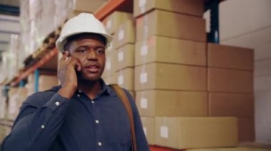 Beyaz kasklı Afrikalı erkek yönetici depoda yürürken cep telefonuyla konuşuyor.