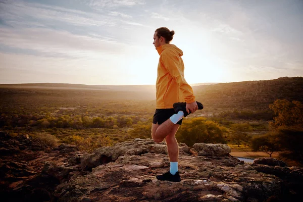 Atleta masculino haciendo ejercicio de estiramiento de piernas preparándose para correr en sendero natural por la mañana con vista al amanecer —  Fotos de Stock