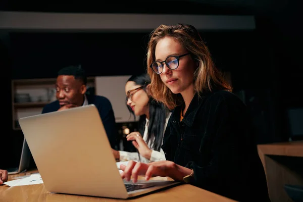 Jovem empresária vestindo óculos sentada com seus colegas no local de trabalho usando laptop à noite — Fotografia de Stock