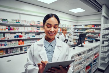 Başarılı genç bayan eczacı eczanede dijital tablet kullanıyor ve meslektaşı arka planda bilgisayar kullanıyor.