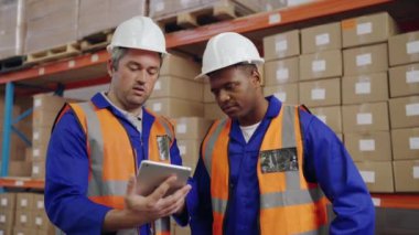 Depodaki siparişleri kontrol ederken dijital tablete bakan üniformalı iki erkek işçinin portresi.