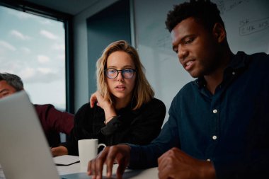 Beyaz ve Afrikalı kadın ve erkek iş arkadaşları ofiste otururken dizüstü bilgisayarla bir şey tartışıyorlar.