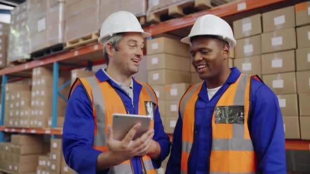 Dva šťastní multietničtí mužští dělníci v záchranné vestě a bílém klobouku smějící se společně při pohledu na digitální tablet ve velkém skladu — Stock video