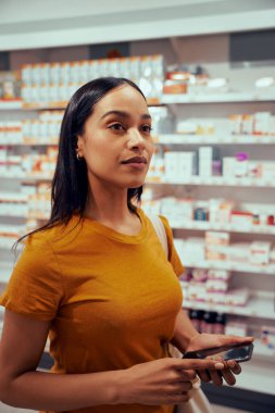 Akıllı telefonlu genç bir kadın eczanede durmuş rafa bakıyor.
