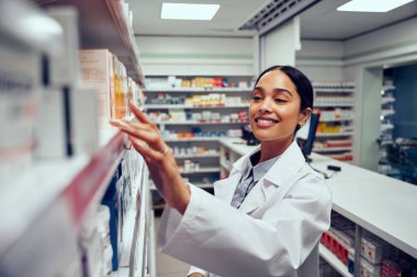 Smiling young female pharmacist wearing labcoat standing behind counter looking for medicine in shelf clipart