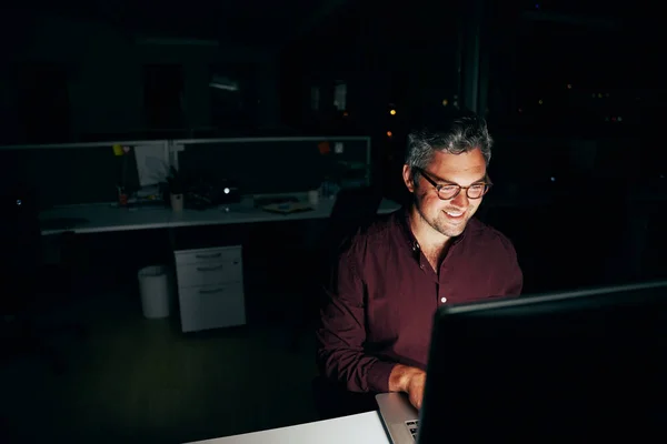 Portret van een lachende zakenman die laat in de avond aan een computer aan zijn bureau werkt. — Stockfoto