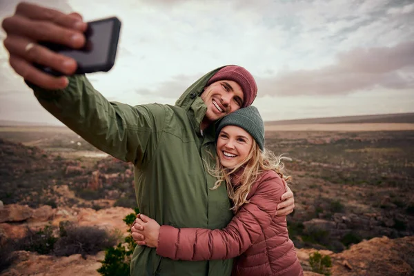 Alegre joven pareja abrazándose durante el campamento de montaña y tomando selfie usando teléfono inteligente contra el fondo de la naturaleza — Foto de Stock