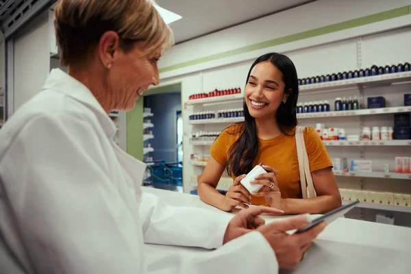 快乐的高级女药剂师在与年轻顾客交谈时，会向他们展示数码平板电脑的显示屏，让他们在药店里拿着一瓶补充剂 — 图库照片