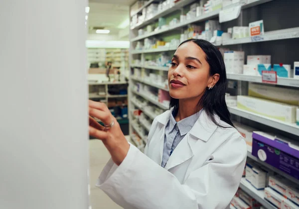 Νεαρή γυναίκα φαρμακοποιός που φοράει παλτό ψάχνοντας για φάρμακα στο ράφι στο φαρμακείο — Φωτογραφία Αρχείου