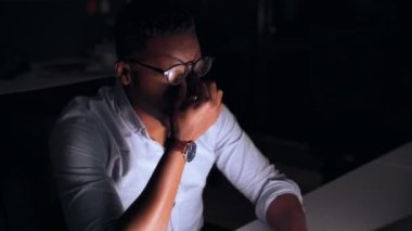 Karma ırk işadamı gece geç saatlere kadar çalışıp, laptopun teslim tarihini yazarken gözlerini ovuşturuyor.