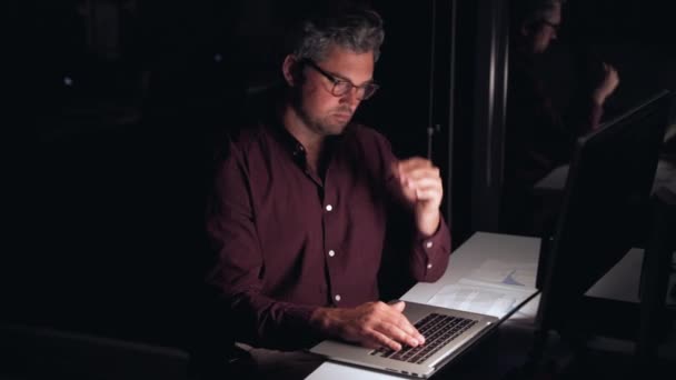 Kaukasisk forretningsmann som jobber sent en kveld og skriver på bærbar PC som strekker seg og hviler med våpen mens han fullfører oppdraget – stockvideo