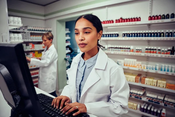 Young woman working in pharmacy typing on computer while checking inventory wearing labcoat standing behind counter