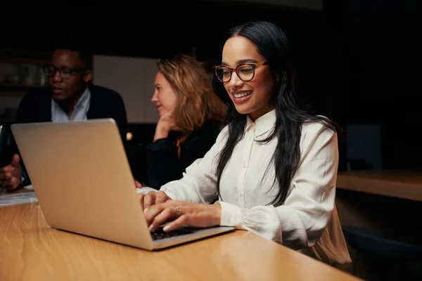 Egy mosolygó fiatal üzletasszony portréja, aki kollégái mellett ül, és laptopon dolgozik az irodában. — Stock Fotó