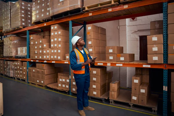 Afrikaanse mannelijke magazijn werknemer met veiligheidsvest en helm werken in groot magazijn met behulp van digitale tablet — Stockfoto