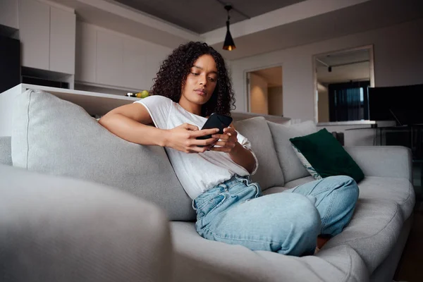 Raça mista bela afro-americana mulher relaxante em casa digitando no dispositivo celular sentado no sofá — Fotografia de Stock