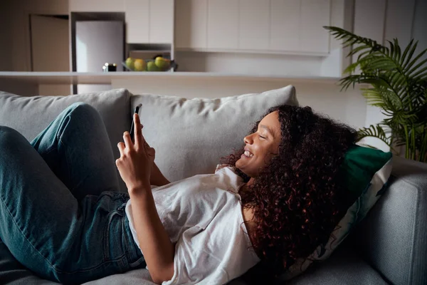 Mezcla de raza femenina adolescente acostado en sofá mensajes de texto en el dispositivo celular — Foto de Stock