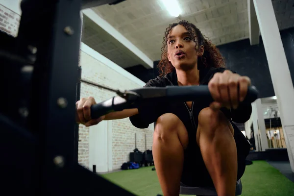 Afro-americana usando uma máquina de remo durante o treinamento cross fit. Atleta feminina se exercitando intensamente no ginásio. Foto de alta qualidade — Fotografia de Stock