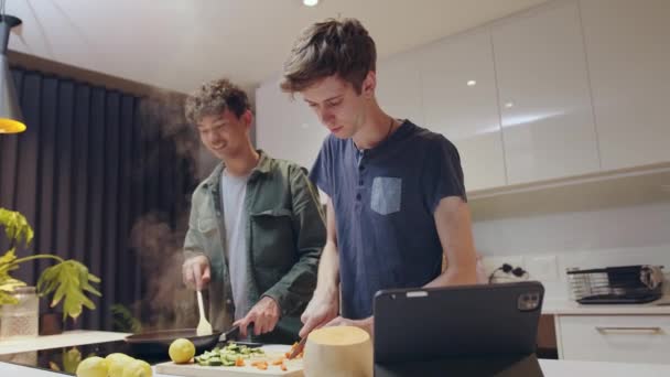 Μικτή φυλή gay ζευγάρι μαγείρεμα μαζί στην κουζίνα χρησιμοποιώντας φορητό υπολογιστή για καθοδήγηση — Αρχείο Βίντεο