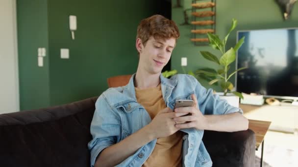 Caucasiano masculino relaxante no sofá mensagens de texto no dispositivo celular em casa — Vídeo de Stock