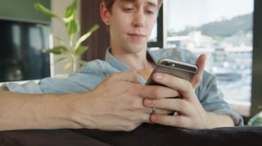 Caucasian male texting on cellular device while relaxing on sofa chatting to friends taking study break 