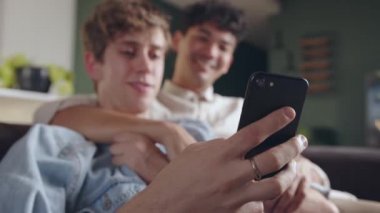Young male gay couple enjoying time together relaxing on couch scrolling on cellular device 