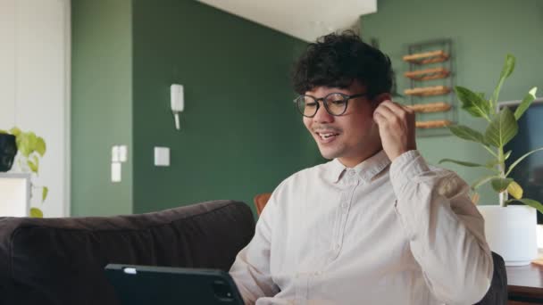 Mixed race male wearing spectacles and listening with earphones while engaging on online call using digital tablet — стоковое видео