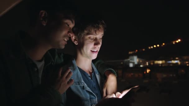 Male gay mixed race coupe standing on balcony together at night scrolling through social media while chatting — стоковое видео