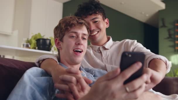 Молодая гей-пара смешанной расы проводит время вместе целуясь на диване, держа в руках сотовое устройство — стоковое видео