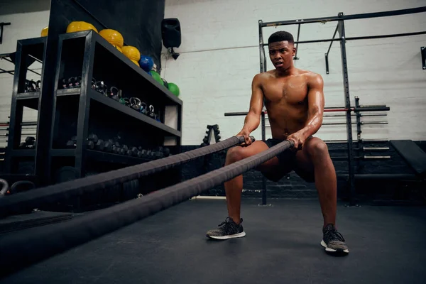 Jovem afro-americano se exercitando dentro de um ginásio. Topless personal trainer masculino usando corda para fortalecer seus braços. Foto de alta qualidade — Fotografia de Stock