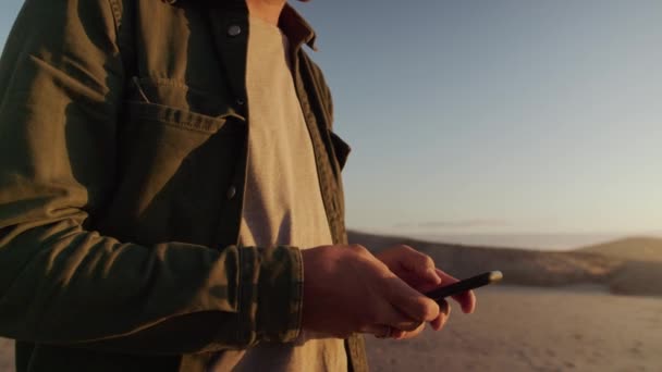 En ung, hvit mann som tekstet på telefonen på stranden. Nære på at mannen ser på telefonen. Opptak av høy kvalitet på 4k – stockvideo