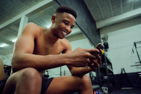 Giovane maschio afroamericano guardando il suo telefono per rispondere a un messaggio online. Felice, maschio razza mista sorridente al suo telefono mentre seduto in palestra. Foto di alta qualità — Foto Stock