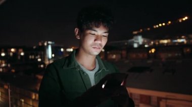 Mixed race male typing on cellular device standing on balcony over looking city lights 