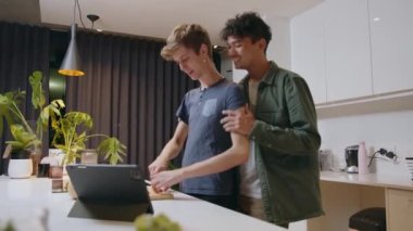 Romantic male couple cooking in kitchen using recipe on digital tablet showing affection