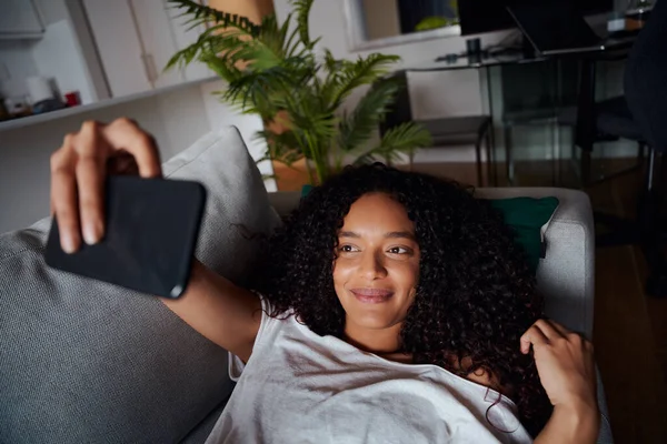Змішана раса жінка бере селфі, лежачи на дивані посміхаючись — стокове фото