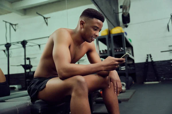 Jovem afro-americano olhando para seu telefone para responder a uma mensagem online. Corrida mista, treinador pessoal masculino olhando para o telefone enquanto se senta no ginásio. Foto de alta qualidade — Fotografia de Stock