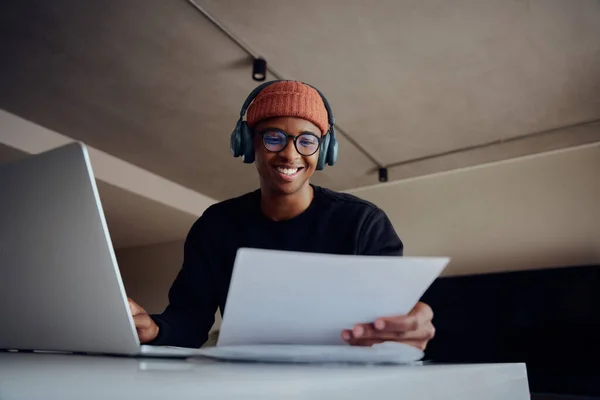 アフリカ系アメリカ人男性はノートパソコンを使って家庭で働いている。幸せなアフリカ系アメリカ人男性が自宅から懸命に仕事をしながら紙を保持。高品質の写真 — ストック写真