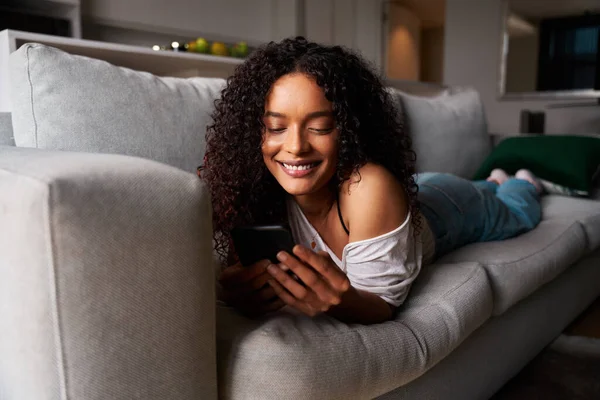 Estudiante de raza mixta relajándose en mensajes de texto de sofá en el dispositivo celular — Foto de Stock