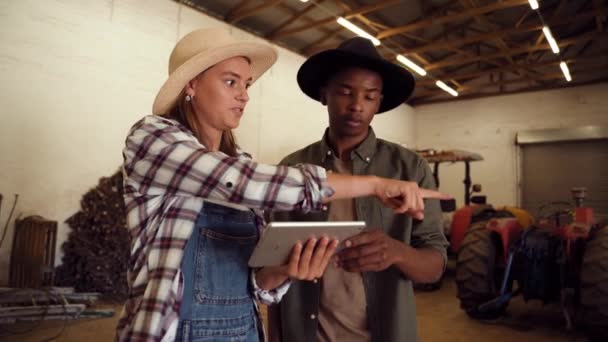 Blanke vrouwelijke boer met digitale tablet, terwijl gemengde ras mannelijke punten rond schuur kamer — Stockvideo