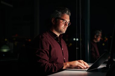 Ciddi bir işadamı pencerenin yanında oturuyor ve gece geç saatlerde ofiste dizüstü bilgisayarla çalışıyor.