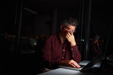 Orta yaşlı bir işadamı dizüstü bilgisayarın önünde gece yarısına kadar proje üzerinde çalışırken gözlerini ovuşturuyor.
