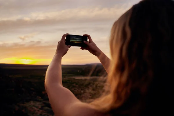 Vista trasera de una mujer joven sosteniendo un teléfono inteligente tomando fotos del amanecer de la mañana contra el horizonte — Foto de Stock