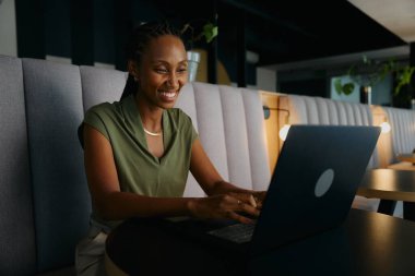 Modern ofis salonundaki masada dizüstü bilgisayar kullanan gülümseyen siyahi iş kadını.