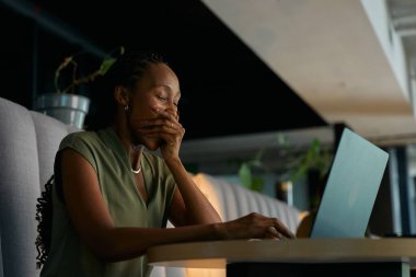 Modern ofis lobisinde dizüstü bilgisayarla çalışırken esnemekten yorgun düşmüş siyahi iş kadını.