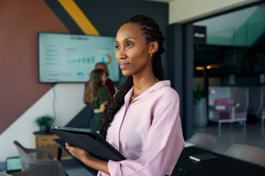 Modern ofiste tabletli düşünceli genç bir iş kadını ve arka planda meslektaşları var.