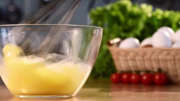 Крупним планом знімок з збивання свіжих сирих курячих яєць у скляній мисці з заливанням молока в процесі. Блискучий барвистий фон з білими яйцями та зеленим салатом . — стокове відео