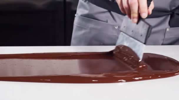 De banketbakker werkt met chocolade. Vrouwelijke kok in grijs uniform temperen gesmolten chocolade op een witte stenen tafel met behulp van een schraper en pyrometer. — Stockvideo