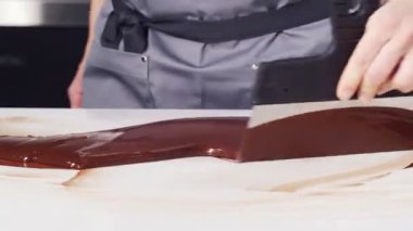 Pasta şefi kadın kazıyıcı ve pirometre kullanarak beyaz taş bir masada erimiş çikolata serpiştiriyor. Şekerci, kahverengi çikolata kaplama.